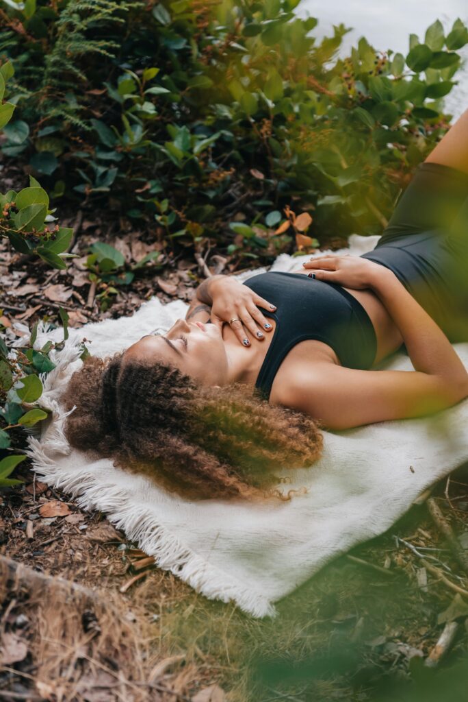 A woman with curly hair meditates on a white towel amidst greenery outdoors, embracing nature.